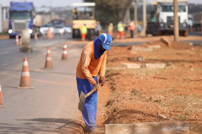 Trabalhador com pá em obra de pavimentação, com cones e outros operários ao fundo